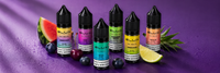 Six 10ml Elux nic salt e-liquid bottles and boxes in different flavour colours arranged with fresh fruit on a deep purple studio background.
